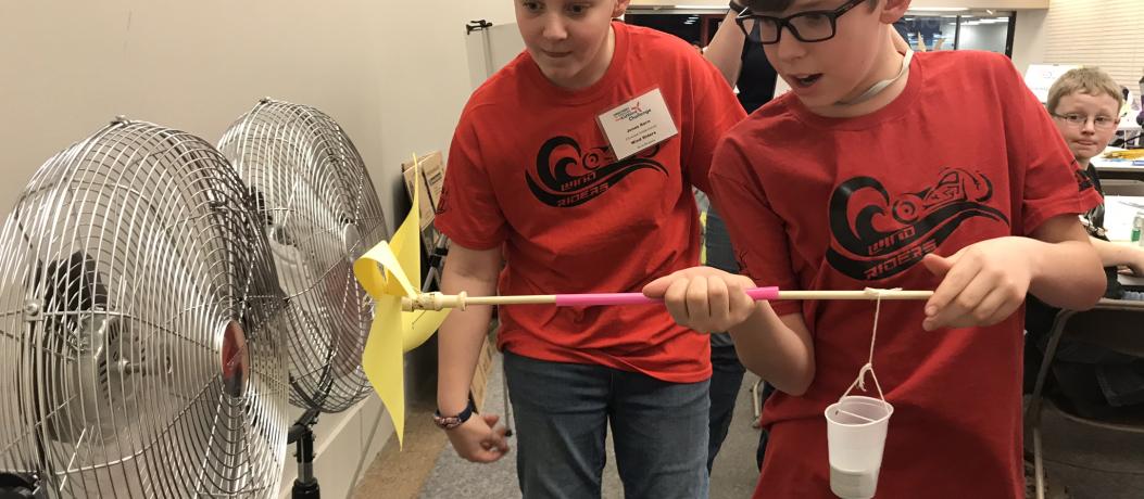 Two boys holding a handheld windmill they made in front of a fun. They are using wind to turn the windmill and lift weights attached on a string.