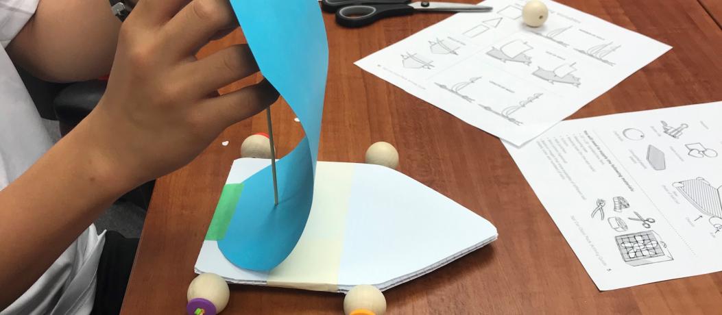 A student's hands are shown as he builds a small sailcar (base is corrugated plastic, wheels are wooden beads, skewers are axels, sail is turqoise construction paper)