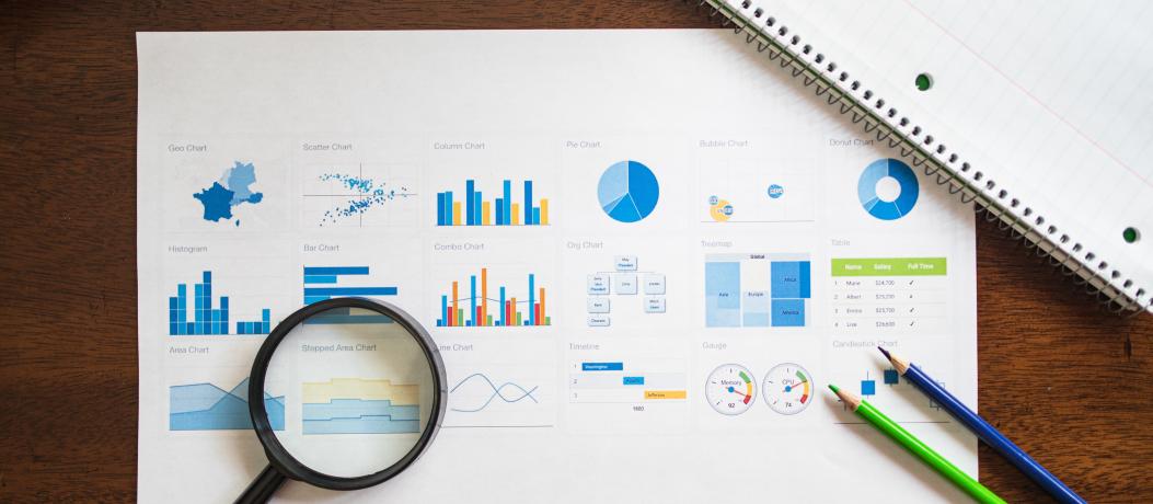 Items laying on desk: Sheet of paper with charts and graphs, notepad, magnifying glass, and pencils