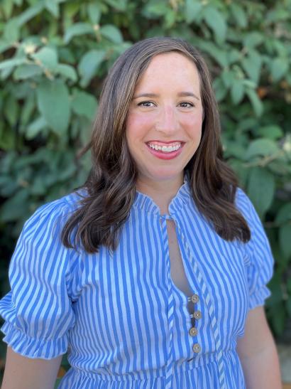 Close-up of woman with brown hair wearing blue and white vertically-striped shirt. Leaves and greenery in background.