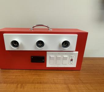 a box on a table that has 3 lightbulbs, 3 switches, a watt meter, and an outlet