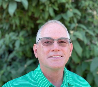 Close-up of man wearing glasses and a green shirt. Leaves and greenery in background.